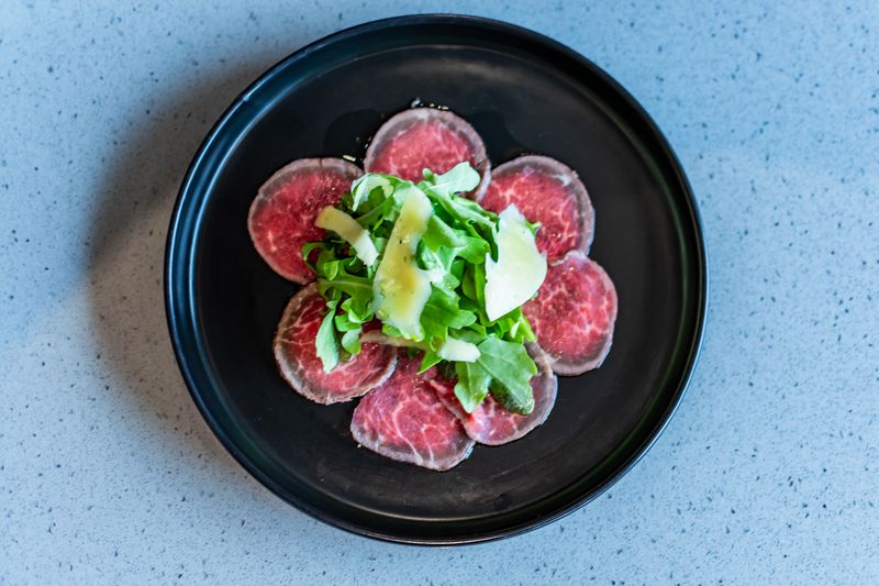 Thinly sliced beef with rocket and shaved cheese on a black plate